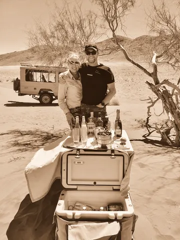 A table laid in desert sand beside a vehicle, bottles set out